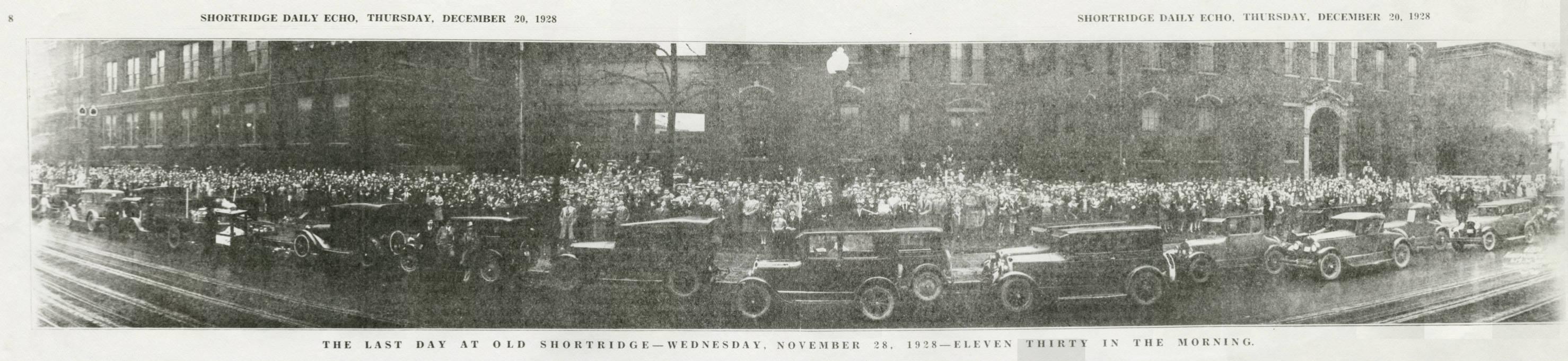 A large crowd of people fill out the yard in front of a large brick school. Cars line the street in front of the crowd.