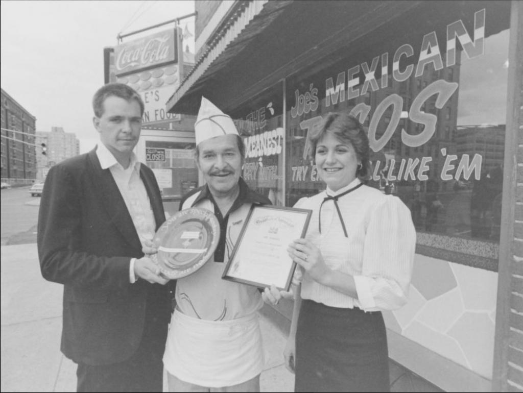 Standing in front of the restaurant, Joe Rangel wears a cook's cap and apron and is flanked by two people who are presenting him an award.