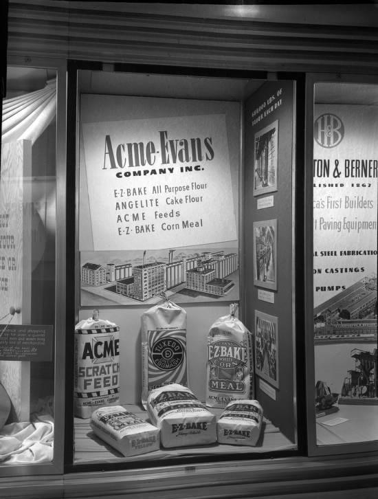 A rectangular window display shows bags containing Acme-Evans products. Behind these are a picture of the Acme-Evans building complex and the company name with a list of their products.