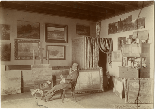 Adams sits in a chair in front of a painting on an easel. The room is filled with paintings hung on the wall and scattered on the floor. 