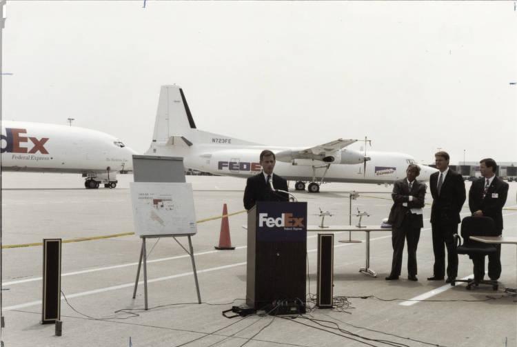 A man stands at a podium and speaks into a microphone on the tarmac at an airport. There are three other mean standing to his right. There is a presentation chart to his left and two FedEx planes behind him.