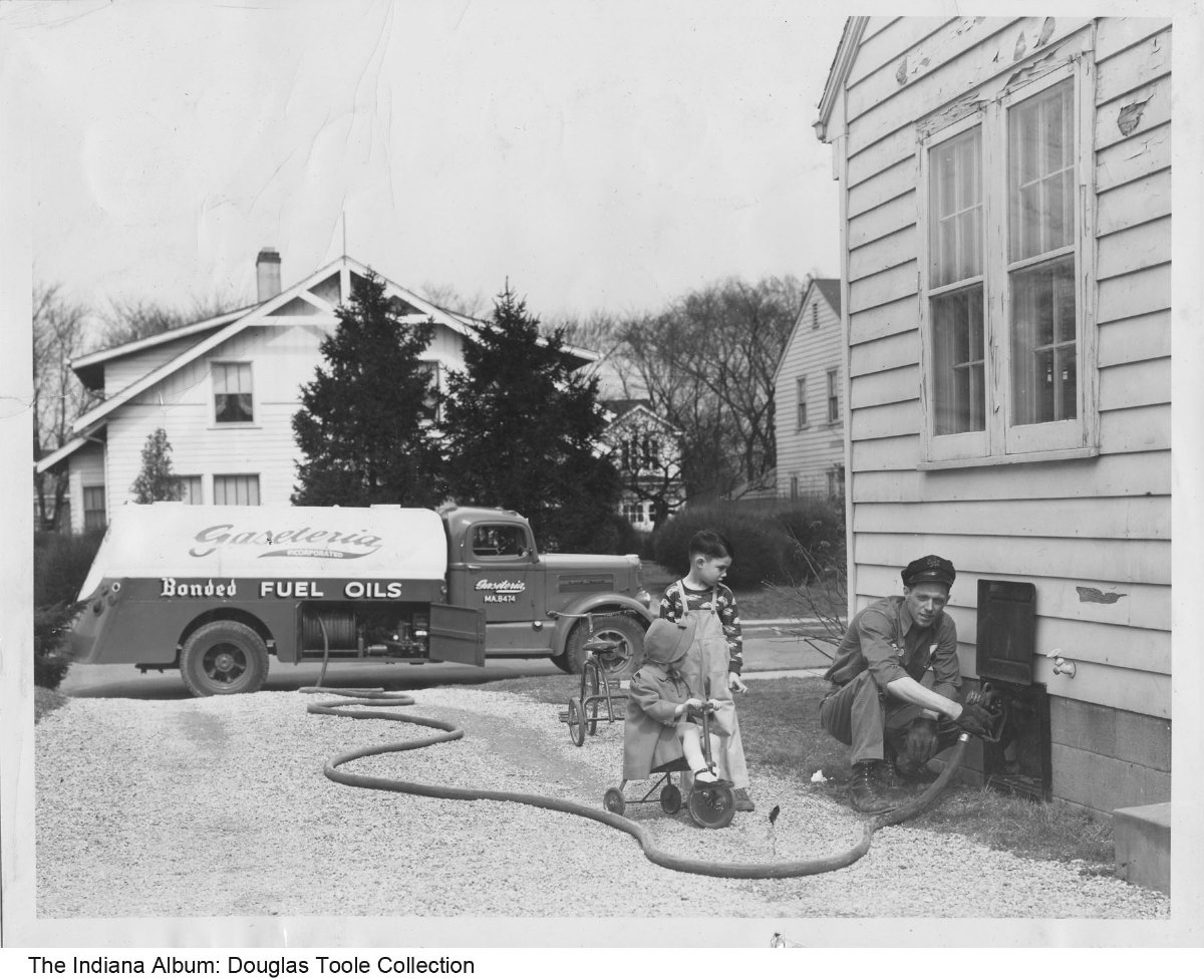 A man pumps oil into a side opening in a house while two children watch. The gas truck is parked at the end of the driveway. 