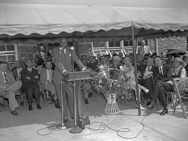 A man stands at a podium speaking. A group of people are seated behind him under an awning. 