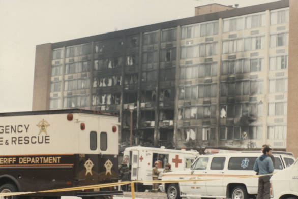 The mainly windowed front of a multi-story building. More than half of the windows are blackened and/or broken, and several emergency vehicles are parked in front.