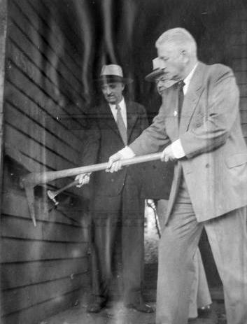 Three men in suits are next to the outside wall of the house. One is using a pickaxe to remove siding and another is using a hammer.