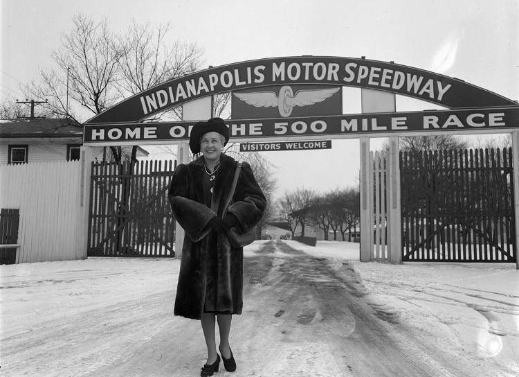 A woman in a fur coat stands in the road under a sign that says "Indianapolis Motor Speedway."