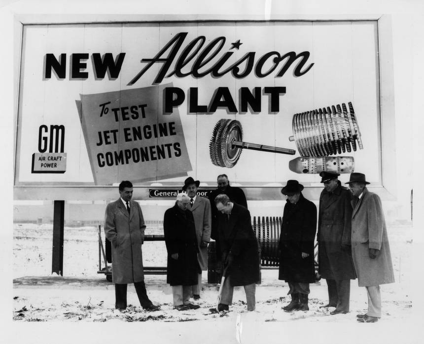 A group of men, one holding a shovel, stand in front of an Allison Engine billboard.