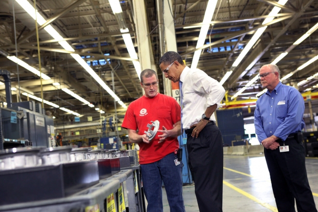 Inside a factory building, a man holds the part. The president is next to him, and another man stands behind the president.