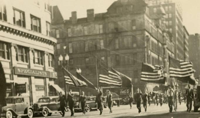 A formation of people, the first two rows of which are holding flags, is in the middle of a street.
