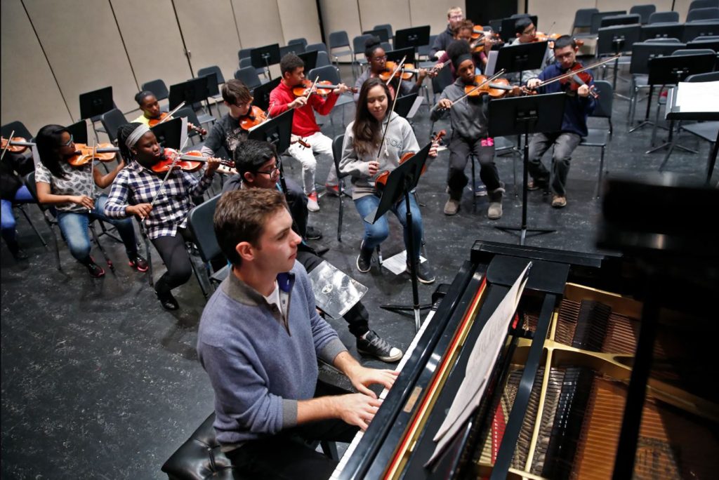 A group of students sit in front of music stands and are playing instruments. A boy plays the piano in the foreground.