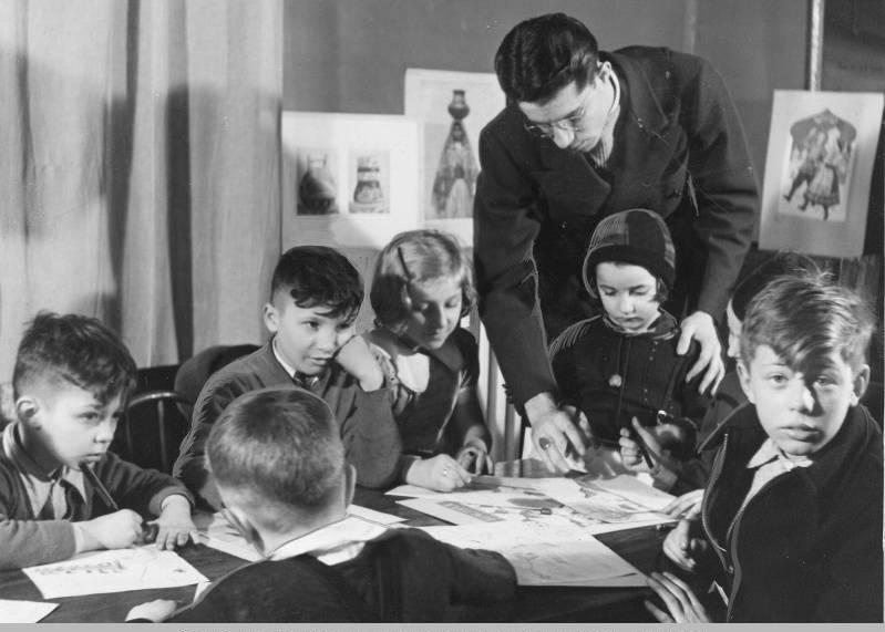 A man instructs a group of children who are sitting at a table drawing.