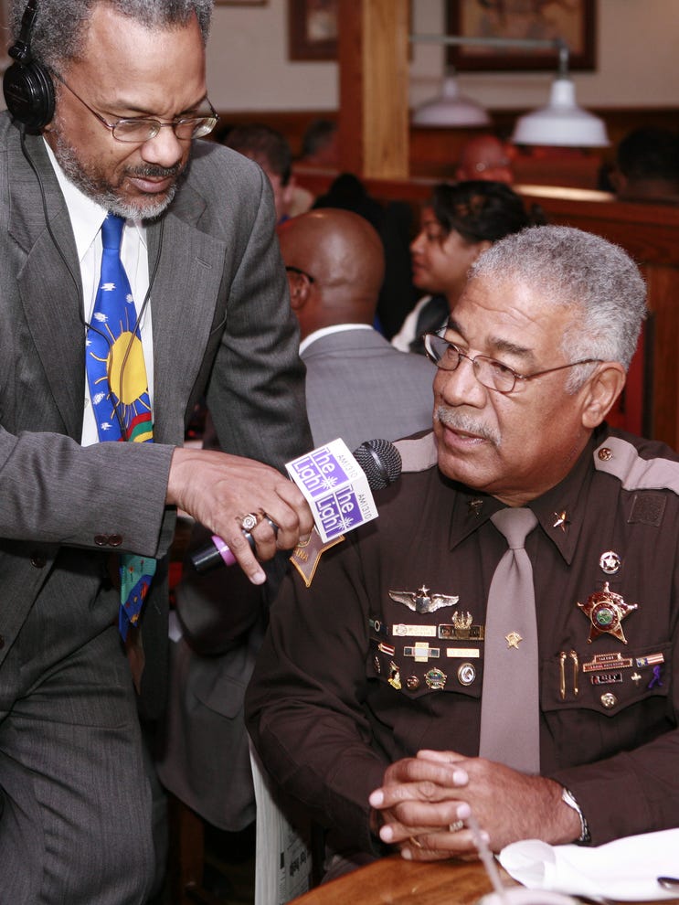 A man wearing a sheriff's uniform and seated at a table is speaking into a microphone being held to him by a man standing beside him.
