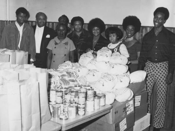 A row of people stands behind tables loaded with turkeys, canned goods and grocery bags.