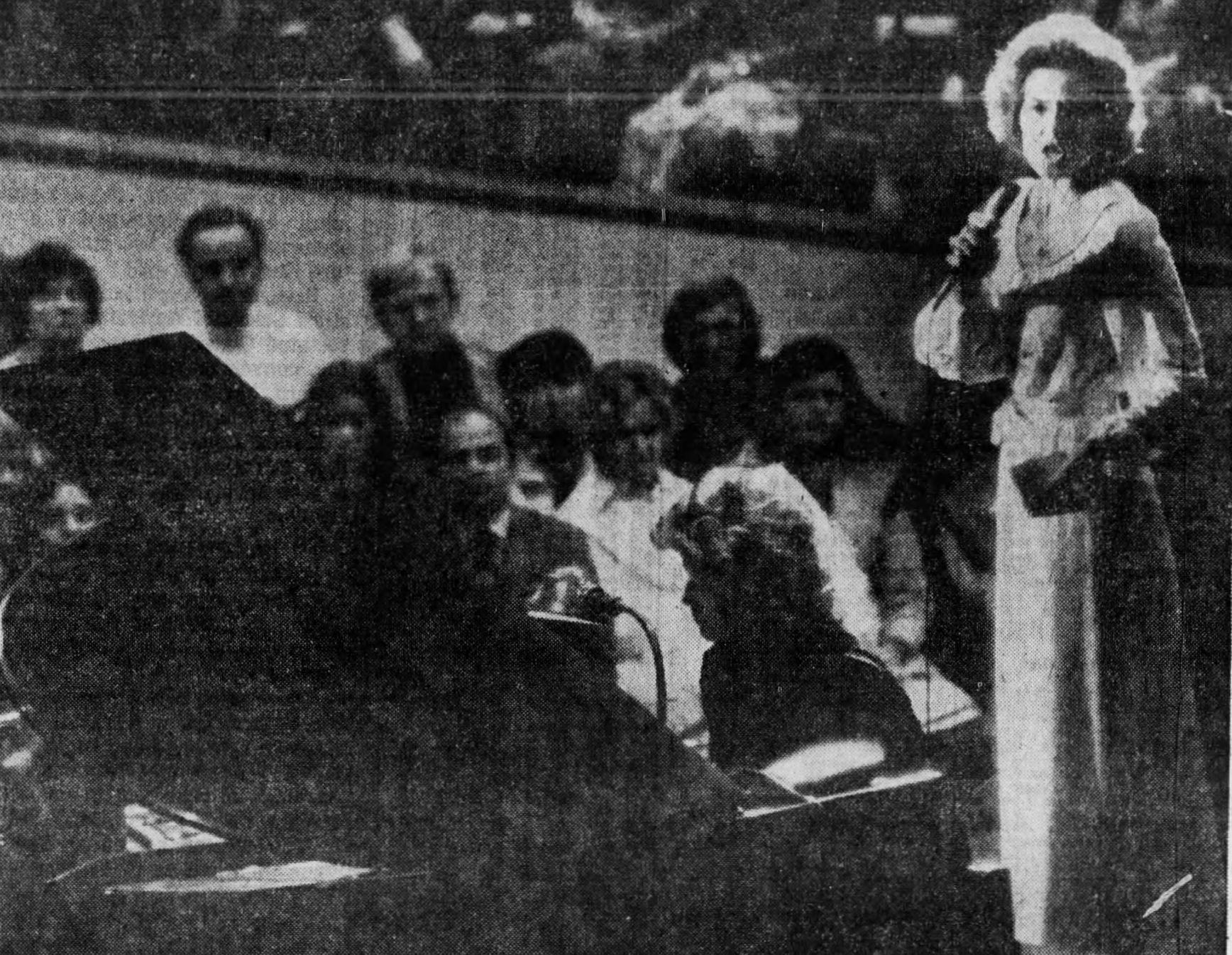 Anita Bryant stands on a stage and speaks into a microphone. A crowd sits in the background. 