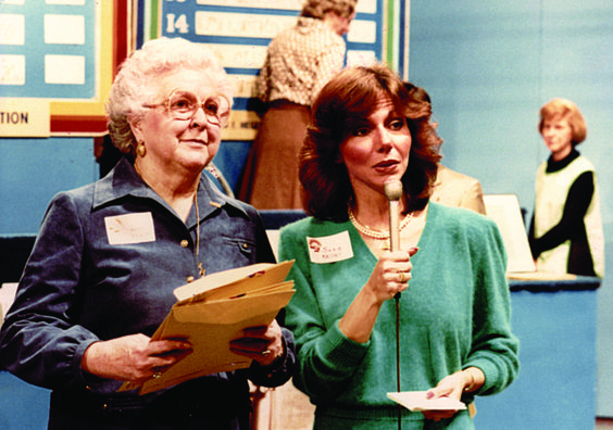Two women are standing in front of a television backdrop. One woman is holding a microphone.