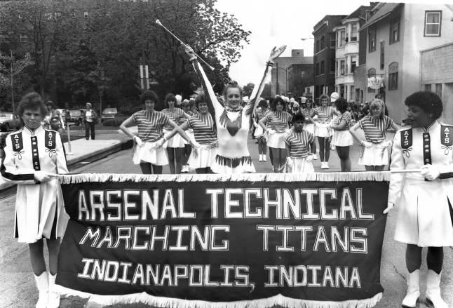 Young women in marching uniforms walk in a parade. Two people are holding a banner that says, "Arsenal Technical Marching Titans."