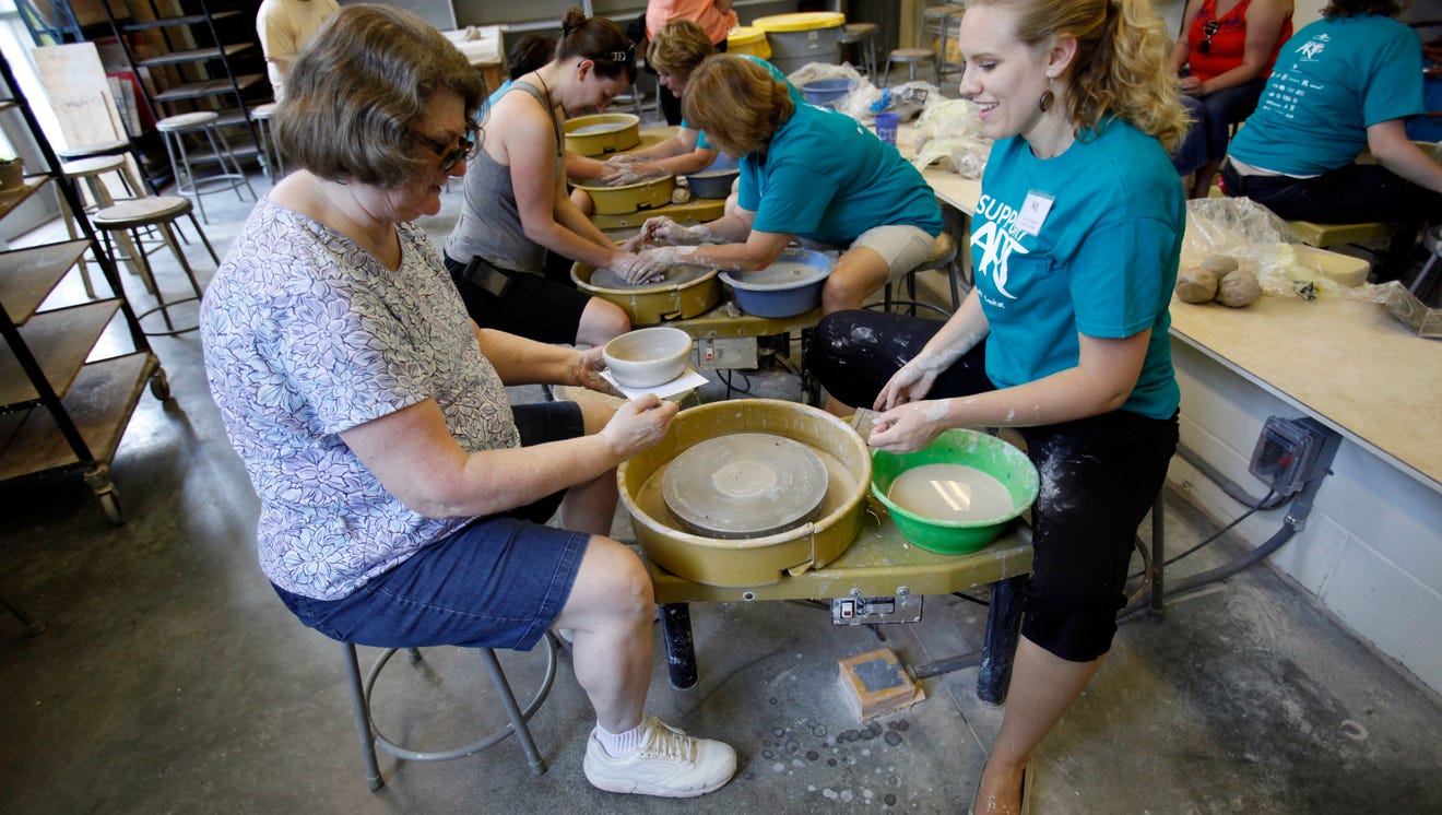 A woman sits on a stool in front of a clay wheel. Another woman in a staff t-shirt sits opposite and helps. 
