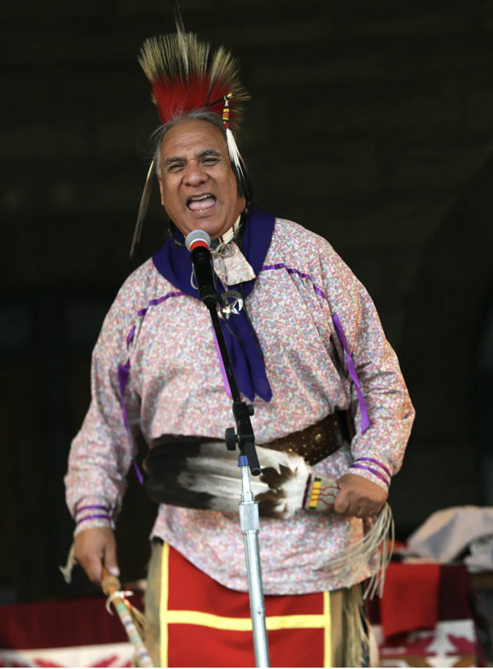A man is dressed in traditional native american dress. He is singing into a microphone. 