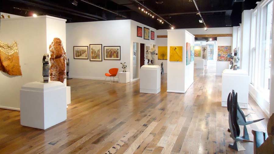 A gallery with natural light coming in from large window. Various types of art is hanging from walls or positioned on free-standing pedestals.
