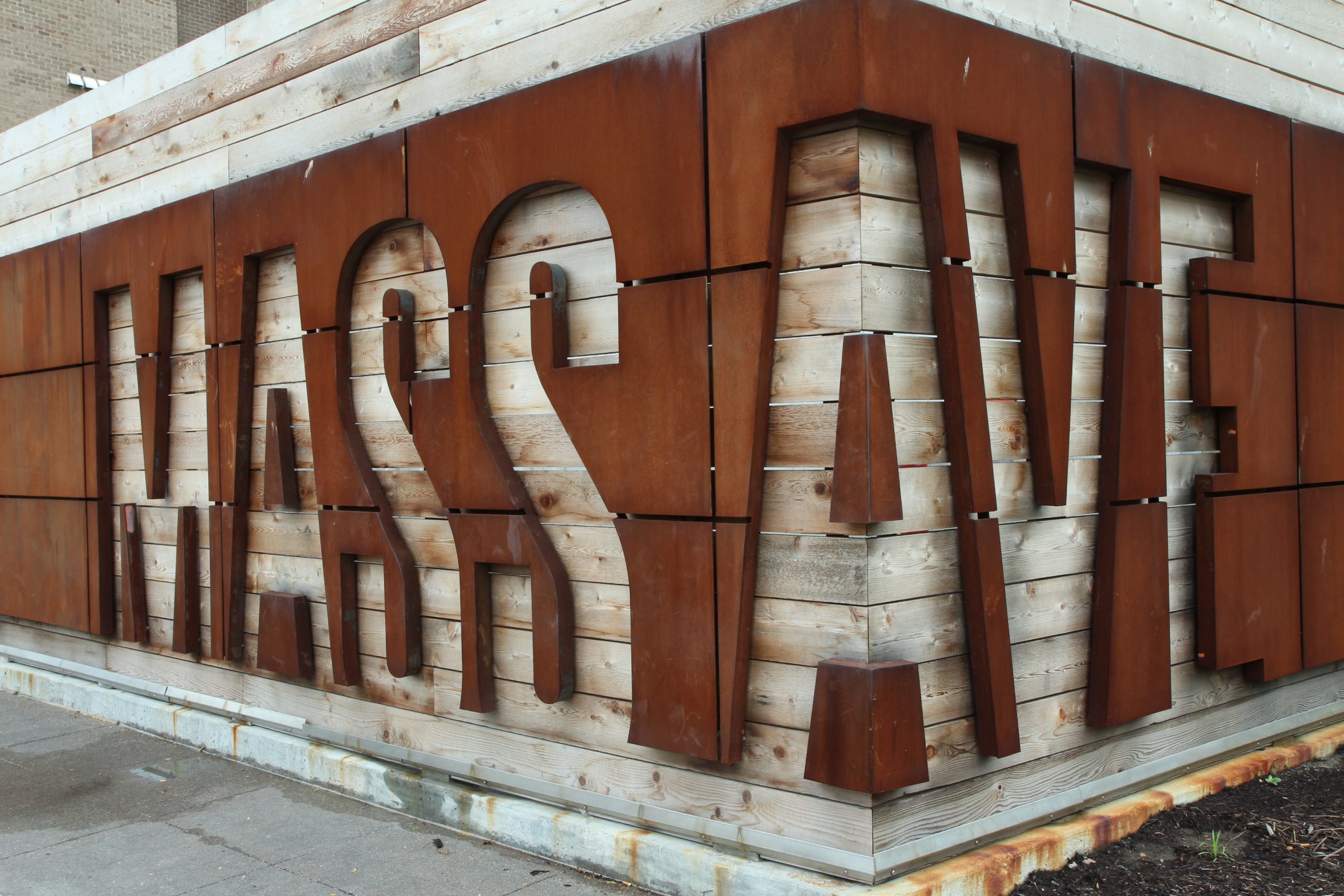 A wood-slat wall with a metal covering. The words "Mass Ave" are cut out of the metal.