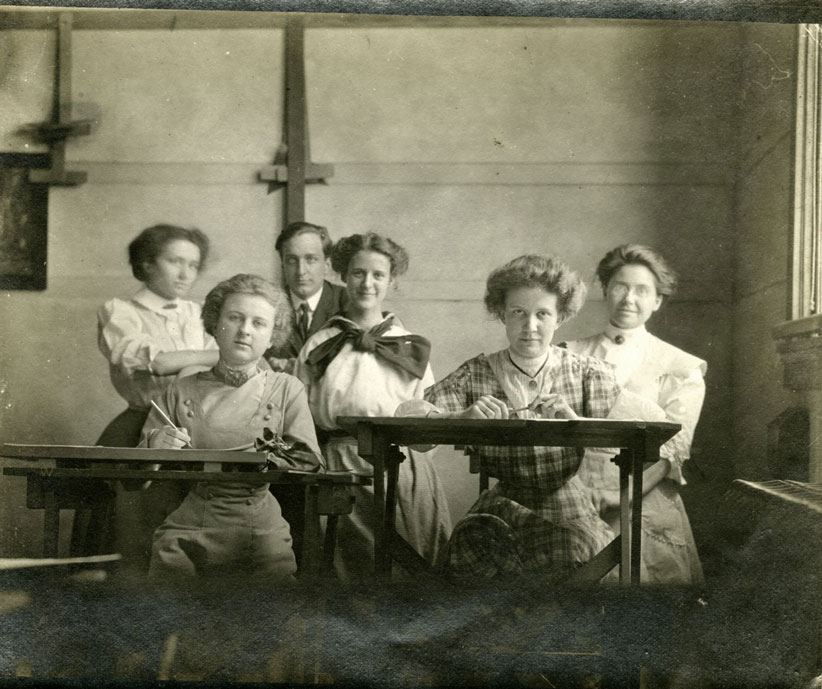 Five women and one man are gathered together. Two women sit at drawing desks. 