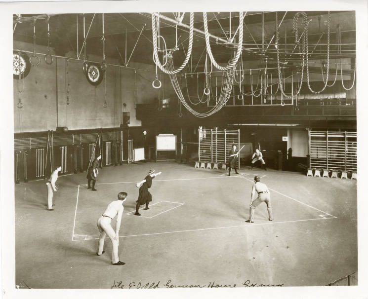 Interior of a large gymnasium showing six men and women playing stickball.