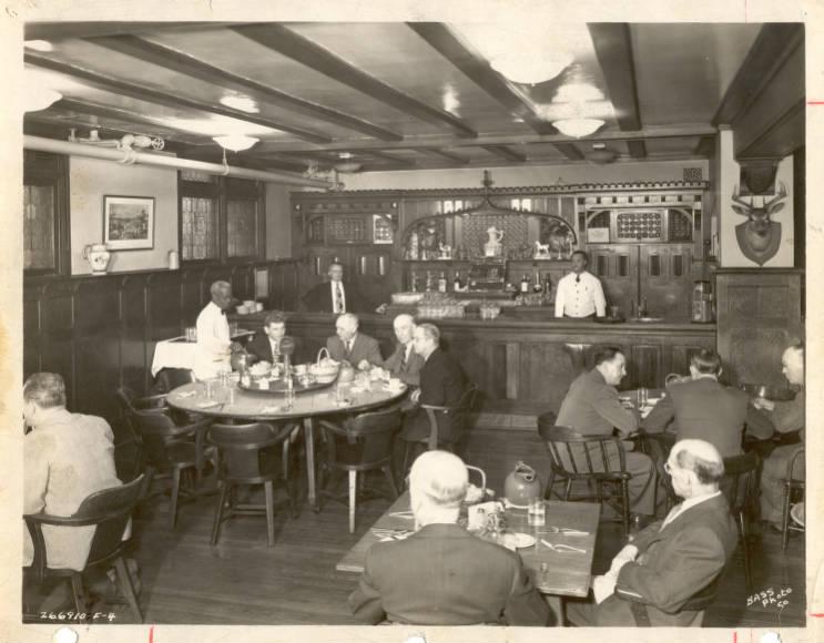 Interior of a wood-paneled restaurant with several people seated at tables and a bar across the back of the room.