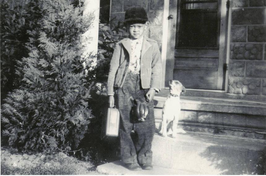 A young child stands at the entrance of a house. A small dog sits behind him.