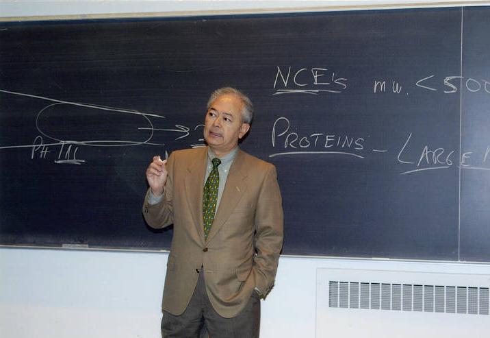 Gus Watanabe stands in front of a chalk board and is giving a lecture.