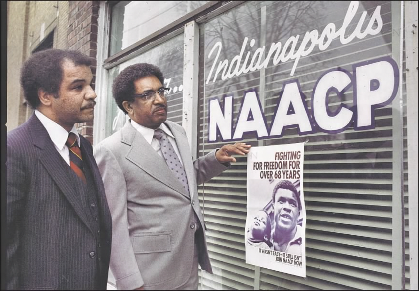 Two men stand outside a building with N A A C P painted on the window.