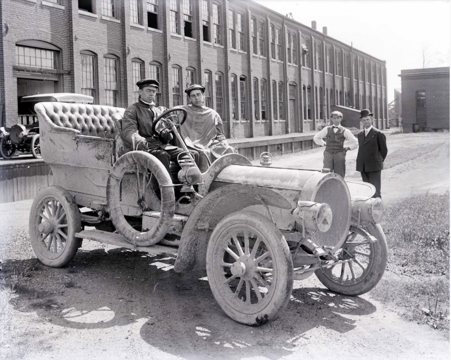 Two men sit in a car. The car is parked in front of a large industrial building. 