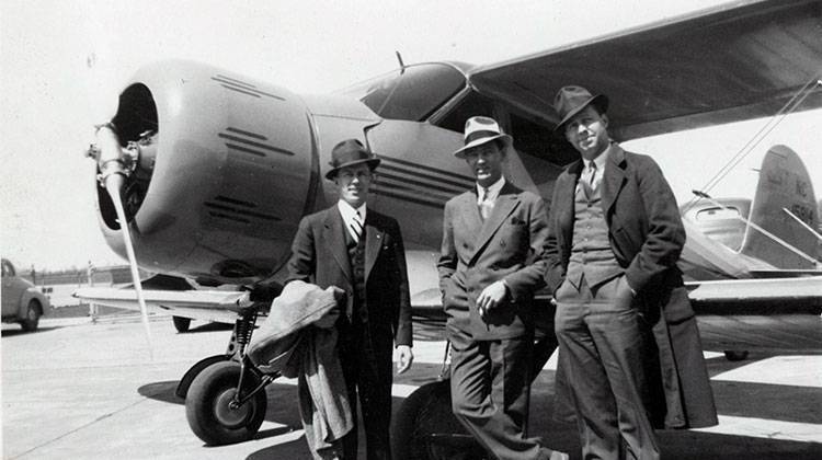 Three very dapper men wearing suits and fedoras pose in front of a biplane.