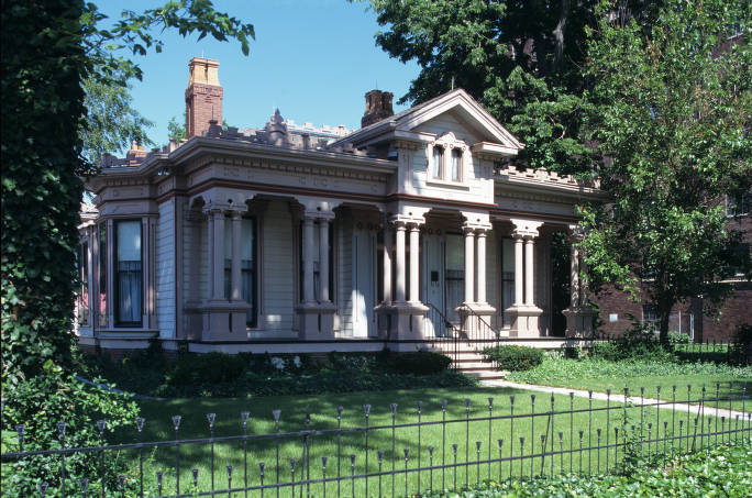 An italiate-style single-story house with six sets of columns on the front porch. A small black iron fence borders the front yard.