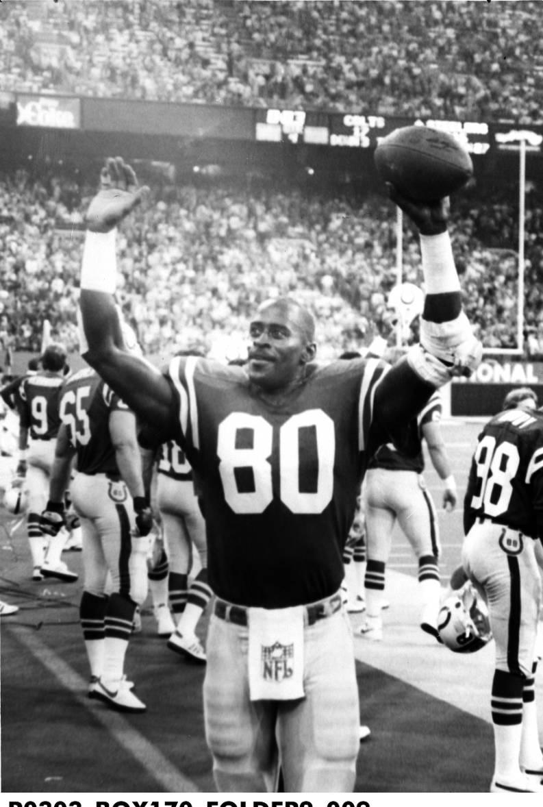 A football player holds up a football over his head. Other players are standing around in the background and the stadium seats are filled with people.