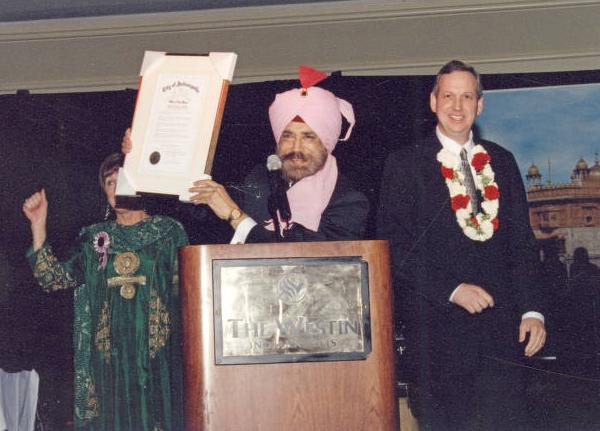 A man stands behind a podium holding up a framed certificate. A woman is on his right, and the mayor is on his left.
