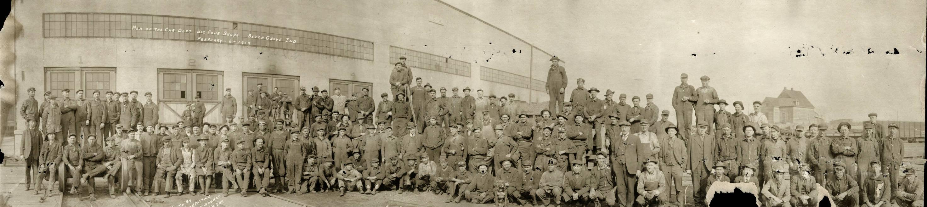 A large group of men stand outside of a factory building. 