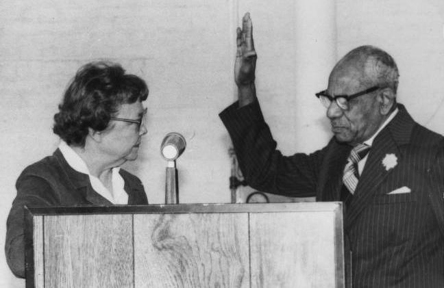 Virginia Dill McCarty and Benjamin A. Osborne stand facing each other behind a podium. Osborne has his hand raised for swearing in. 