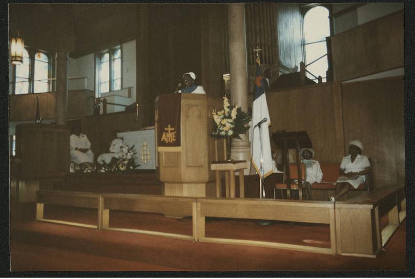 Interior view of the front of a church. A woman is standing at the pulpit. Four other women are arranged in seats behind the pulpit.