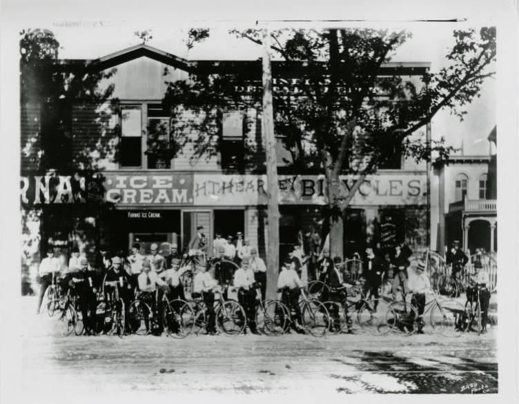 Several men with bicycles line the street and sidewalk in front of a store.