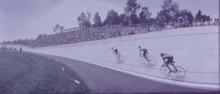 Three cyclists are racing on an outdoor track which is lined on the right by bleachers full of people.