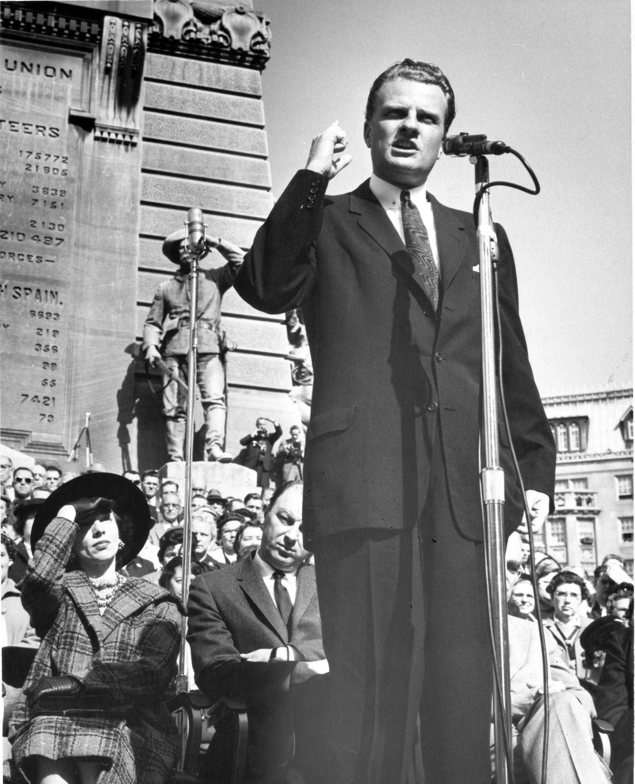 A man stands behind a microphone. A crowd of people are standing around him. A monument is in the background.