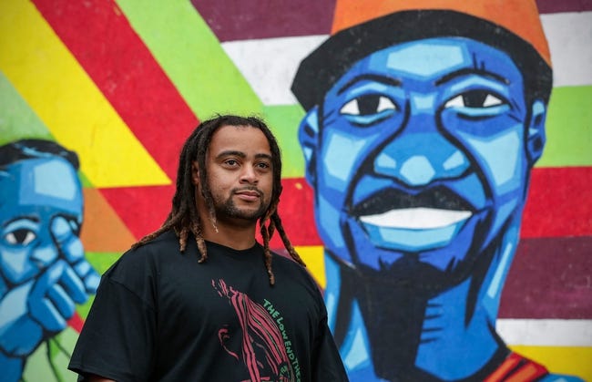 Artist Israel Solomon stands in front of a mural he painted 2020. The painting shows two faces painted in blue against a colorful graphic background.