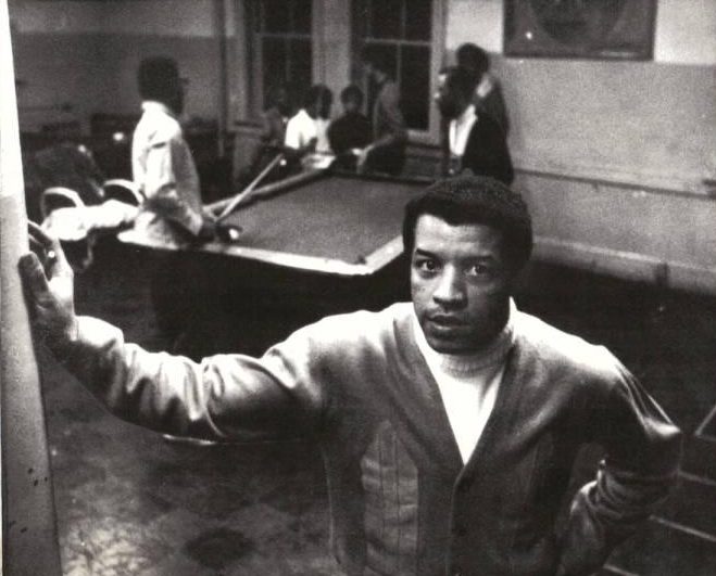 A man stands with his hand on a wall. Men are playing pool in the background.