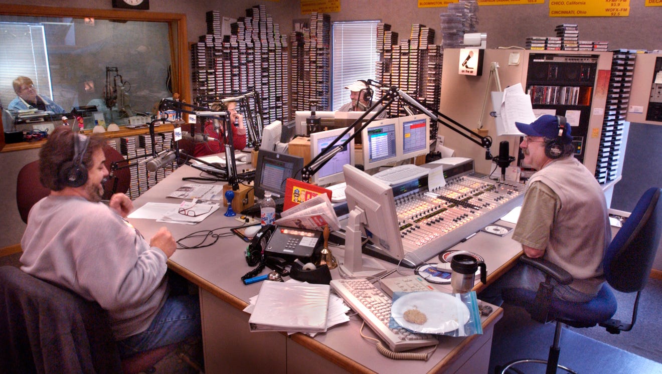Interior of the recording studio with Bob, Tom, Christy Lee and another man doing a live show. They sit around a large desk on which are computer screens, microphones and a sound board.
