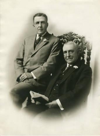 One man is seated in and the other man sits on the arm of a richly carved wooden chair.