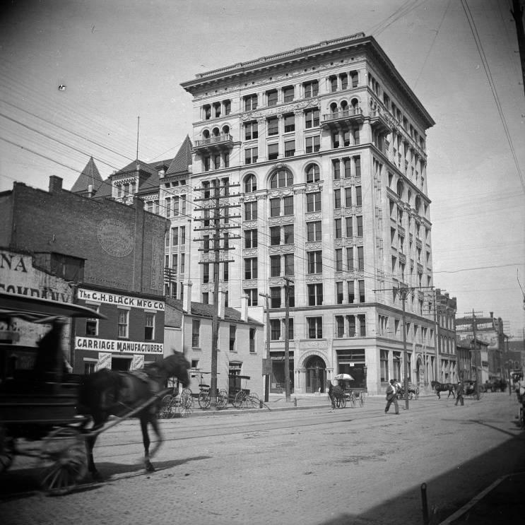 The tall, limestone building is shown in a line of smaller buildings in a street level view which includes people and horse drawn carriages on a wide avenue.