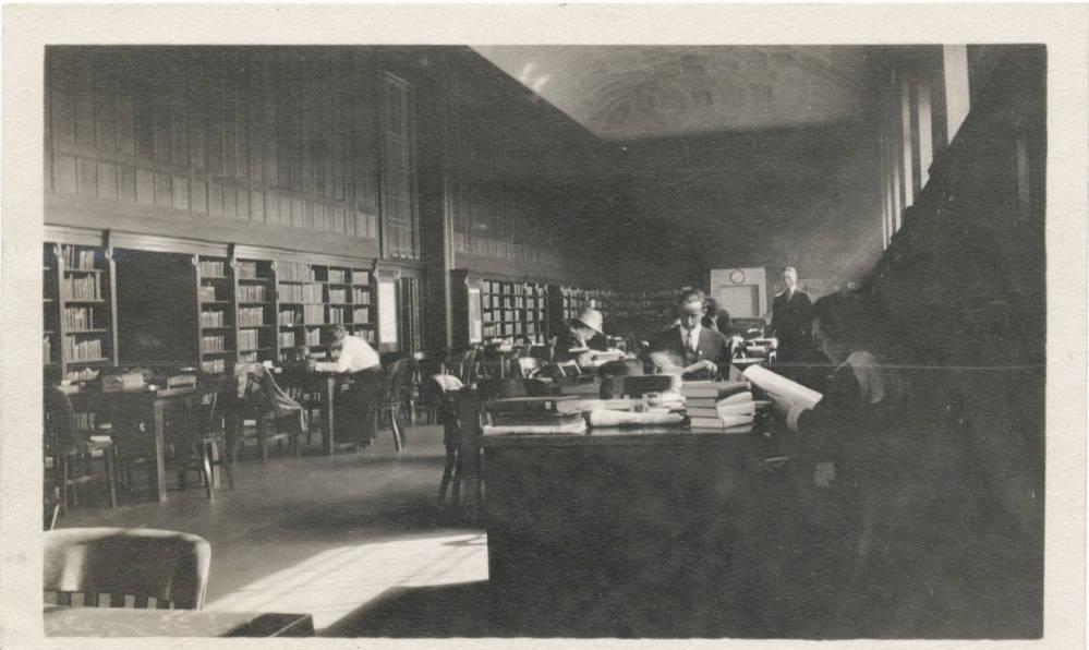 A large room is lined by bookshelves. People sit at tables throughout the room.