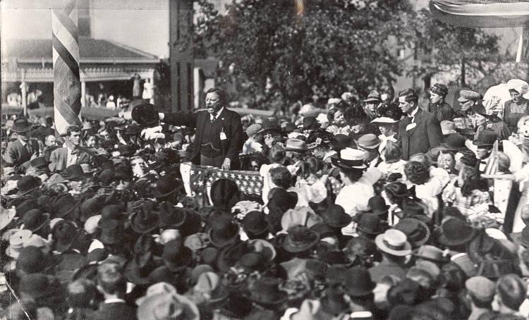 President Theodore Roosevelt stands on a raised dais in the middle of a large crowd of people.