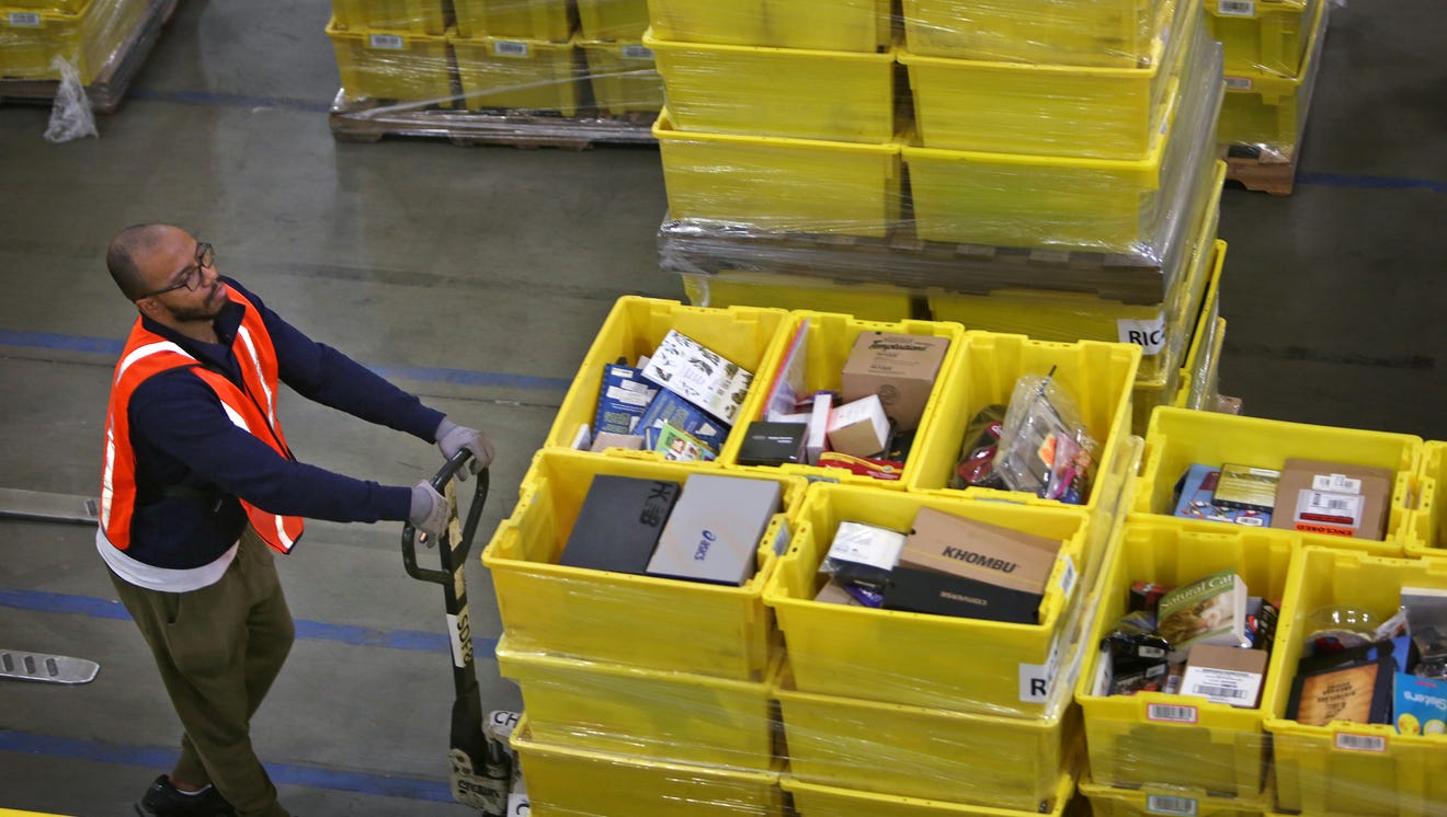 A man wearing a safety vest is pushing a flat card loaded with yellow bins in which are several products.