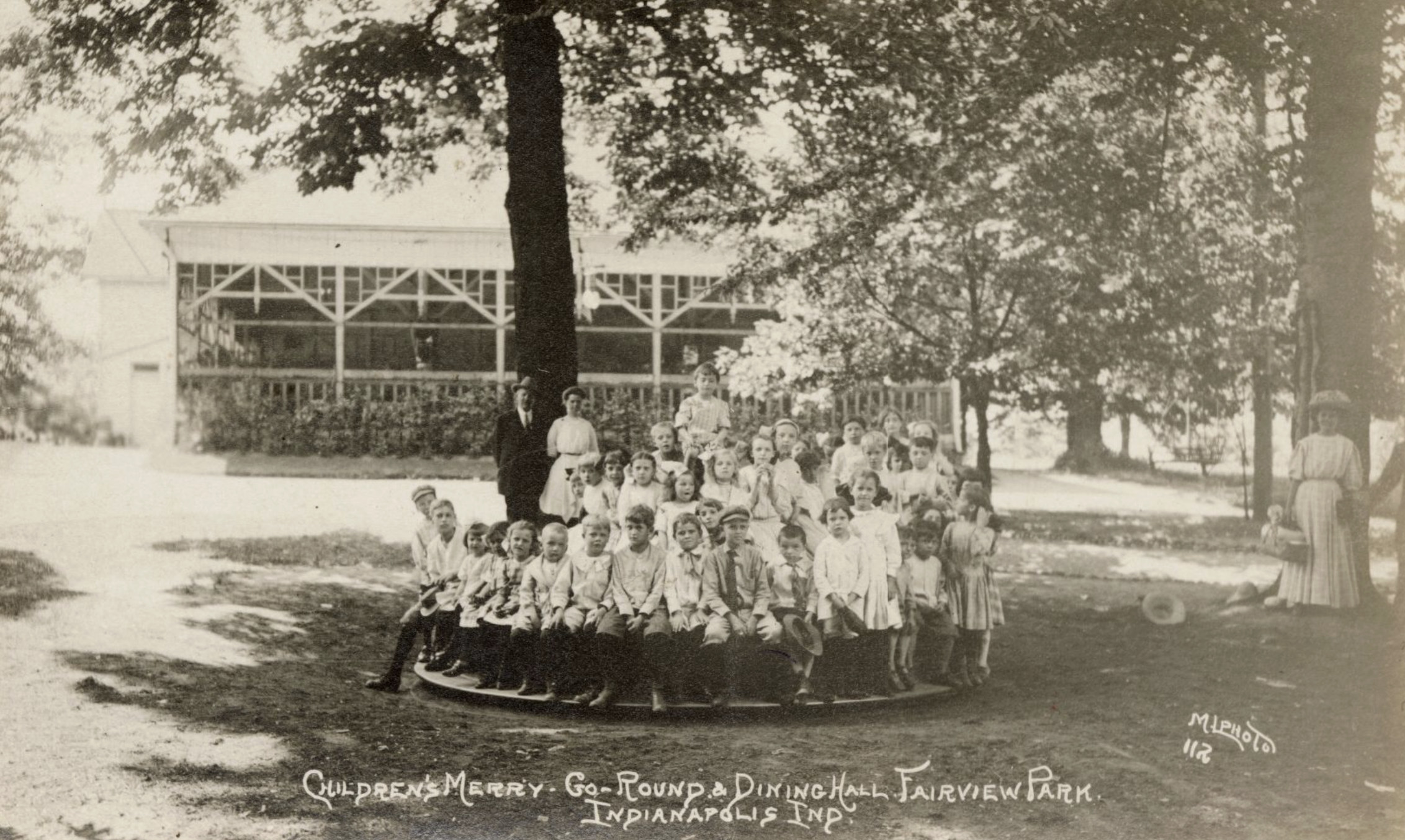 Children sit on the merry-go-round in Fairview Park while adults stand behind them and off to the side.  The park's dining hall stands in the background.